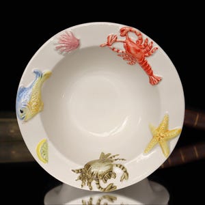 May include: White ceramic bowl with embossed sea life designs. Features a red lobster, yellow starfish, blue and yellow fish, crab, pink seashell, and a lemon slice. A decorative piece for serving or display.