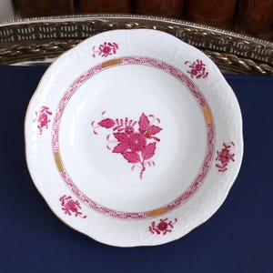 May include: White porcelain bowl with a scalloped edge, featuring a pink floral design in the center and around the rim. The rim has a pink and gold border. The bowl is set on a dark blue surface.