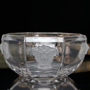 Puede incluir: Un cuenco de cristal transparente y facetado con un diseño de cabeza de Medusa. El cuenco tiene una forma ancha y poco profunda, probablemente para fines decorativos. El cuenco de cristal transparente tiene una superficie reflectante.
