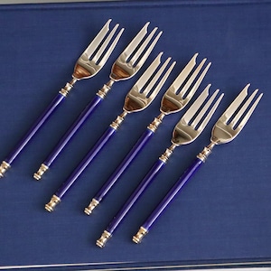 Puede incluir: Seis tenedores plateados con un diseño de tres púas. Los mangos son de color azul oscuro con detalles dorados. Los tenedores están colocados sobre una superficie azul oscuro.