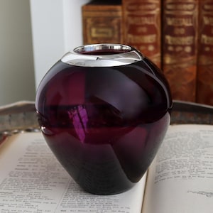 Peut inclure: Un vase rond en verre violet fonc&eacute; avec un bord argent&eacute;. Le vase est pos&eacute; sur un livre ouvert. L'arri&egrave;re-plan comprend une pile de livres avec des lettres dor&eacute;es.