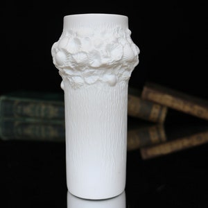 Puede incluir: Jarrón de cerámica blanca con superficie texturizada y diseño floral en relieve alrededor de la parte superior.