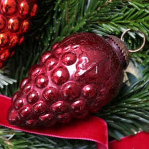 Puede incluir: Un adorno navideño de vidrio rojo oscuro con forma de racimo de uvas. El adorno tiene un acabado brillante y reflectante y está decorado con un lazo dorado metálico.