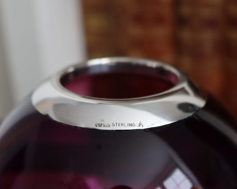 Peut inclure: Gros plan sur un vase en verre violet fonc&eacute; avec un bord argent&eacute;. Le vase a une forme arrondie et une surface lisse et brillante. Le texte "925 STERLING" est estampill&eacute; sur le bord argent&eacute;.