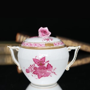 May include: White porcelain sugar bowl with a pink floral design and gold trim. The lid is topped with a pink rose. The bowl has two handles and is a classic piece of tableware.