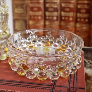 May include: A clear glass bowl with a textured, bubble-like design. The bowl has a rounded shape and is decorated with amber-coloured accents. The bowl is sitting on a red surface, with a stack of similar bowls in the background.
