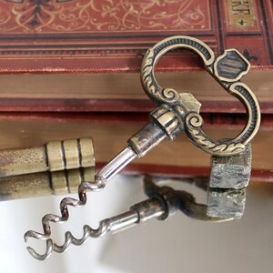 May include: A vintage brass key with a corkscrew attached to the end. The key has an ornate design and is resting on a book with a red leather cover.