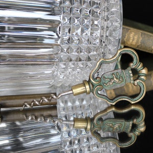 May include: An antique brass and green corkscrew with a decorative handle featuring a crest design. The corkscrew is positioned in front of a textured glass bottle, reflecting on a glossy surface. The overall theme is vintage barware.
