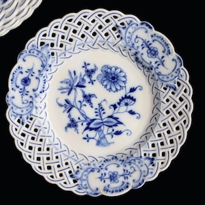 Könnte beinhalten: Weiße Porzellanteller mit blauem Blumenmuster in der Mitte und einem Gitterrand. Der Teller hat einen gewellten Rand und gehört zu einem Set. Das Design ist ein klassisches blau-weißes Muster, ca. 20 cm Durchmesser.