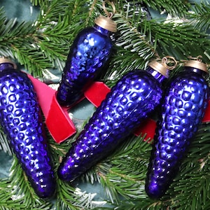 Könnte beinhalten: Vier kobaltblaue Weihnachtsornamente aus Glas in Form von Tannenzapfen. Jedes Ornament hat eine strukturierte Oberfläche und eine goldfarbene Metallkappe. Die Ornamente sind auf einem Bett aus grünen Tannennadeln und rotem Samtband angeordnet.