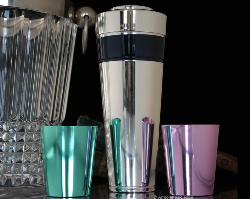 K&ouml;nnte beinhalten: Ein silberner Cocktailshaker mit einem schwarzen Band, zwei Schnapsgl&auml;ser, eines gr&uuml;n und eines rosa, auf einer Holzoberfl&auml;che.