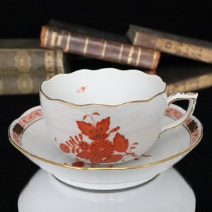 May include: A white porcelain teacup and saucer with a delicate floral pattern in shades of orange and red. The cup has a scalloped rim and a gold trim. The saucer has a matching floral design and a gold rim.