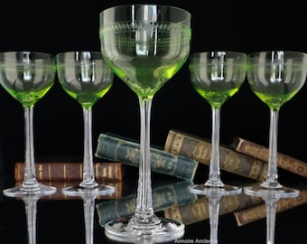 May include: Set of six green glass stemmed wine glasses with a decorative etched pattern around the rim. The glasses are arranged in a row on a dark surface with books behind them.