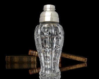 May include: A clear glass cocktail shaker with a silver top. The shaker has a cut glass design and is sitting on a stack of books.