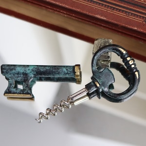 May include: A green and gold key-shaped bottle opener with a corkscrew. The opener has a decorative handle and a small, flat base.