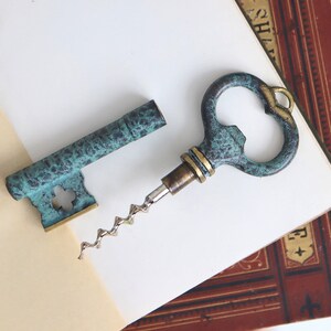May include: A key-shaped bottle opener with a teal and brass finish. The corkscrew is made of silver-coloured metal. The opener is next to a matching key-shaped piece, both resting on a book.