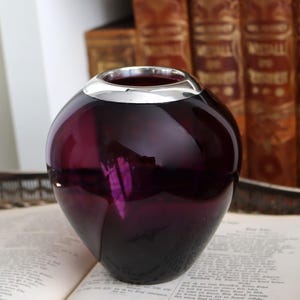 Peut inclure: Un vase sph&eacute;rique en verre violet fonc&eacute; avec un bord argent&eacute;. Le vase est pos&eacute; sur un livre ouvert, avec un arri&egrave;re-plan flou de livres anciens.