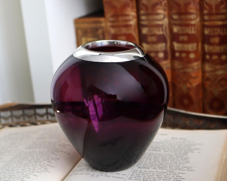 Peut inclure: Un vase sph&eacute;rique en verre violet fonc&eacute; avec un bord argent&eacute;. Le vase est pos&eacute; sur un livre ouvert, avec un arri&egrave;re-plan flou de livres anciens.