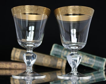 May include: Two clear crystal wine glasses with gold rims and decorative detailing. The glasses have a faceted stem and a wide base. The background is dark, with blurred books visible.