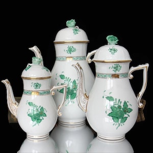 以下が含まれることがあります： 緑色の花柄と金のアクセントが施された、3つの磁器製のコーヒーポット。ポットはサイズが異なり、黒い背景に一列に並んでいます。