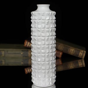 Puede incluir: Un jarrón alto de cerámica blanca con un patrón geométrico texturizado. El jarrón tiene un cuello estrecho y una base ancha. El fondo es negro, con un reflejo del jarrón y algunos libros antiguos.