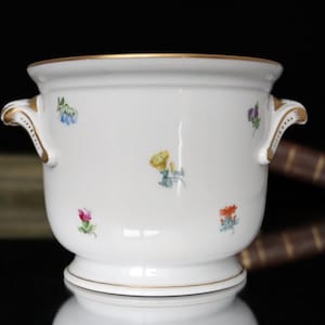 Könnte beinhalten: Ein weißer Porzellanblumentopf mit goldenen Verzierungen und Griffen. Der Topf ist mit kleinen, farbenfrohen Blumenmustern verziert. Die Griffe haben ein dekoratives Golddesign. Der Topf hat einen Sockel.