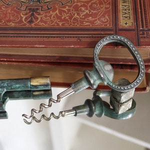Könnte beinhalten: Ein antiker, mehrteiliger Korkenzieher in Grün und Gold mit einem dekorativen, schlüsselförmigen Griff. Der Korkenzieher liegt auf einer reflektierenden Oberfläche mit einem Stapel alter Bücher im Hintergrund.