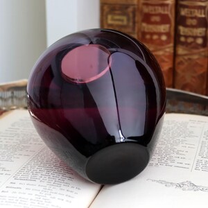 Peut inclure: Un vase en verre violet fonc&eacute; de forme arrondie avec une ouverture circulaire. Le vase est pos&eacute; sur un livre ouvert avec du texte. L'arri&egrave;re-plan comprend des livres anciens. Le vase a une finition brillante et une base sombre.