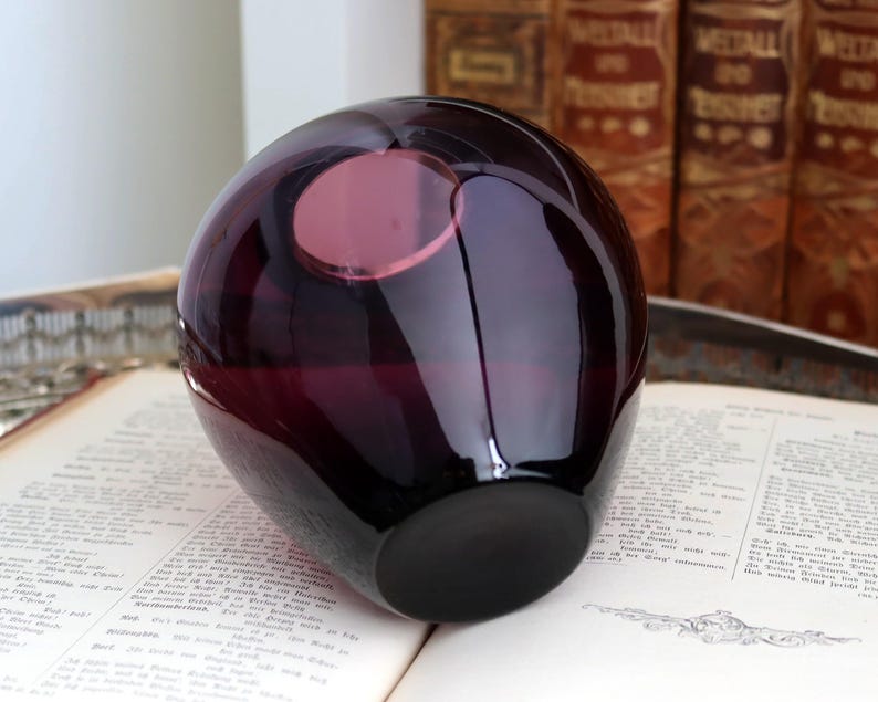 Peut inclure: Un vase en verre violet fonc&eacute; de forme arrondie avec une ouverture circulaire. Le vase est pos&eacute; sur un livre ouvert avec du texte. L'arri&egrave;re-plan comprend des livres anciens. Le vase a une finition brillante et une base sombre.