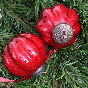 May include: Two glossy red Christmas baubles with ribbed detailing and antique-style metal caps. One bauble has a decorative metal cap. The baubles rest on a bed of green pine needles, with a red ribbon visible.