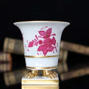 May include: A white porcelain vase with a pink floral design and gold accents. The vase has four gold lion paw feet and sits on a gold base.