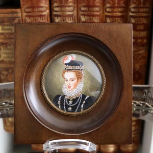 以下が含まれることがあります： 黒いドレスを着て白いフリルカラーと宝石のネックレスをつけた女性の額装されたミニチュア肖像画。肖像画は、正方形の木製フレーム内の丸いフレームに収められています。背景は柔らかくニュートラルな色です。