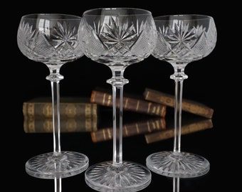 May include: Three clear glass champagne coupes with intricate cut glass designs. The glasses are arranged in a row on a black surface with a stack of books behind them.