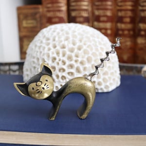 May include: A vintage cat-shaped corkscrew with a brass body and a black head. The corkscrew is in a playful pose, with its tail forming the screw. The cat is set against a blue surface and a white coral-like object.
