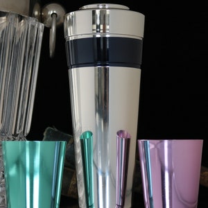 K&ouml;nnte beinhalten: Ein silberner Cocktailshaker mit einem schwarzen Band um die Mitte. Vor dem Shaker stehen drei kleine Metallbecher, einer ist t&uuml;rkis, einer rosa und einer hellgr&uuml;n.