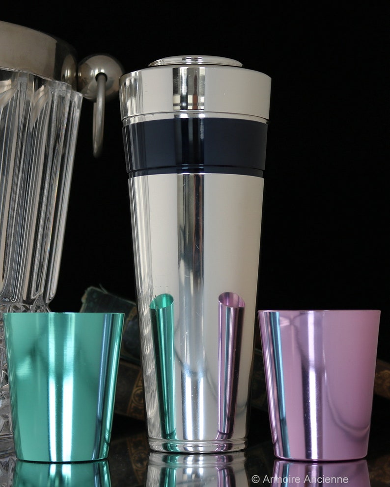 K&ouml;nnte beinhalten: Ein silberner Cocktailshaker mit einem schwarzen Band um die Mitte. Vor dem Shaker stehen drei kleine Metallbecher, einer ist t&uuml;rkis, einer rosa und einer hellgr&uuml;n.