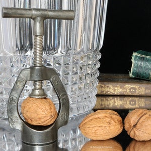 Peut inclure: Un casse-noisette en métal vintage avec une poignée en bois et une vis en métal. Le casse-noisette est ouvert et une noix est dans les mâchoires. Deux noix sont sur une surface sombre.