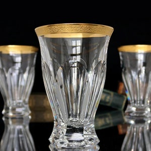 May include: Three clear glass tumblers with a gold rim and a cut glass design. The glasses are sitting on a black surface.