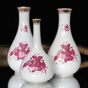 May include: Three white porcelain vases with pink floral designs and gold accents. The vases are of different sizes and are arranged in a row.