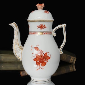 Puede incluir: Una cafetera de porcelana blanca con un diseño floral en tonos naranja y dorado. La cafetera tiene una tapa con un pequeño pomo floral y un pico curvo. La cafetera está decorada con un patrón floral en el cuerpo y el asa.