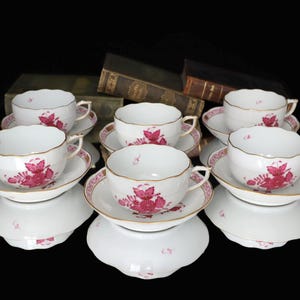May include: A set of six white teacups and saucers. Each cup and saucer features a pink floral design with gold trim. The cups and saucers are arranged on a reflective surface, with antique books in the background.