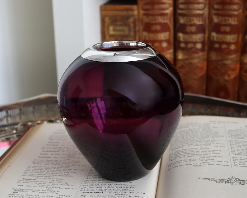 Peut inclure: Un vase rond en verre violet fonc&eacute; avec un bord argent&eacute;. Le vase est pos&eacute; sur un livre ouvert. L'arri&egrave;re-plan comprend une pile de livres avec des lettres dor&eacute;es.