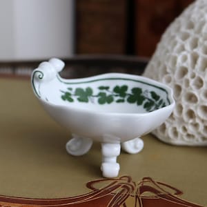 May include: A small, white porcelain dish with three feet and a decorative handle. The rim is adorned with a green floral pattern. The dish is likely for decorative purposes.