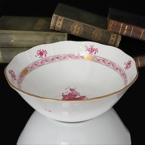 May include: A white porcelain bowl with a scalloped rim, featuring a pink floral and gold border design. The bowl has a gold rim and a matching floral pattern inside. Antique books are in the background.