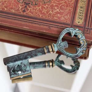May include: An antique-style, ornate key with a green patina finish and gold accents. The key is resting on top of a closed book with a red cover and gold lettering that reads "E.S. Werke".