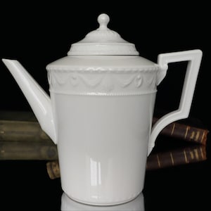 May include: A white porcelain teapot with a lid and a square handle. The teapot has a spout and is decorated with a detailed embossed pattern around the middle. The background is black.
