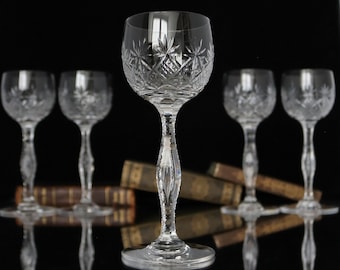 May include: Set of six clear crystal stemmed glasses with intricate cut designs. The glasses are arranged in a row with a stack of books in the background.