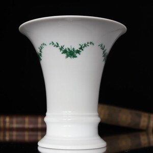 May include: A white porcelain vase with a green floral design. The vase has a flared top and a round base.