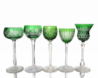 May include: Five green crystal wine glasses with detailed cut patterns. The glasses have clear stems and bases. The green bowls of the glasses are the focal point, with intricate cuts that catch the light. The glasses are of varying shapes.