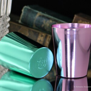 K&ouml;nnte beinhalten: Zwei metallische Becher, einer gr&uuml;n und einer rosa, mit gl&auml;nzender, reflektierender Oberfl&auml;che. Die Becher sind aus Aluminium und haben ein schlichtes, zylindrisches Design.